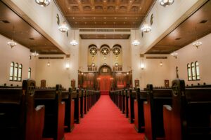 Interior of a synagogue with wooden pews, red carpet aisle, and ornate ceiling with hanging lights.