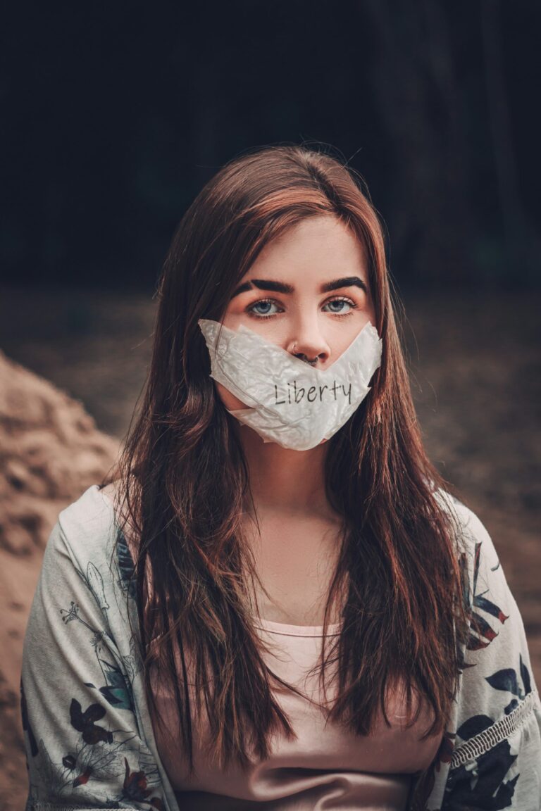 Woman with long brown hair wears a cloth labeled Liberty covering her mouth, sitting outdoors.