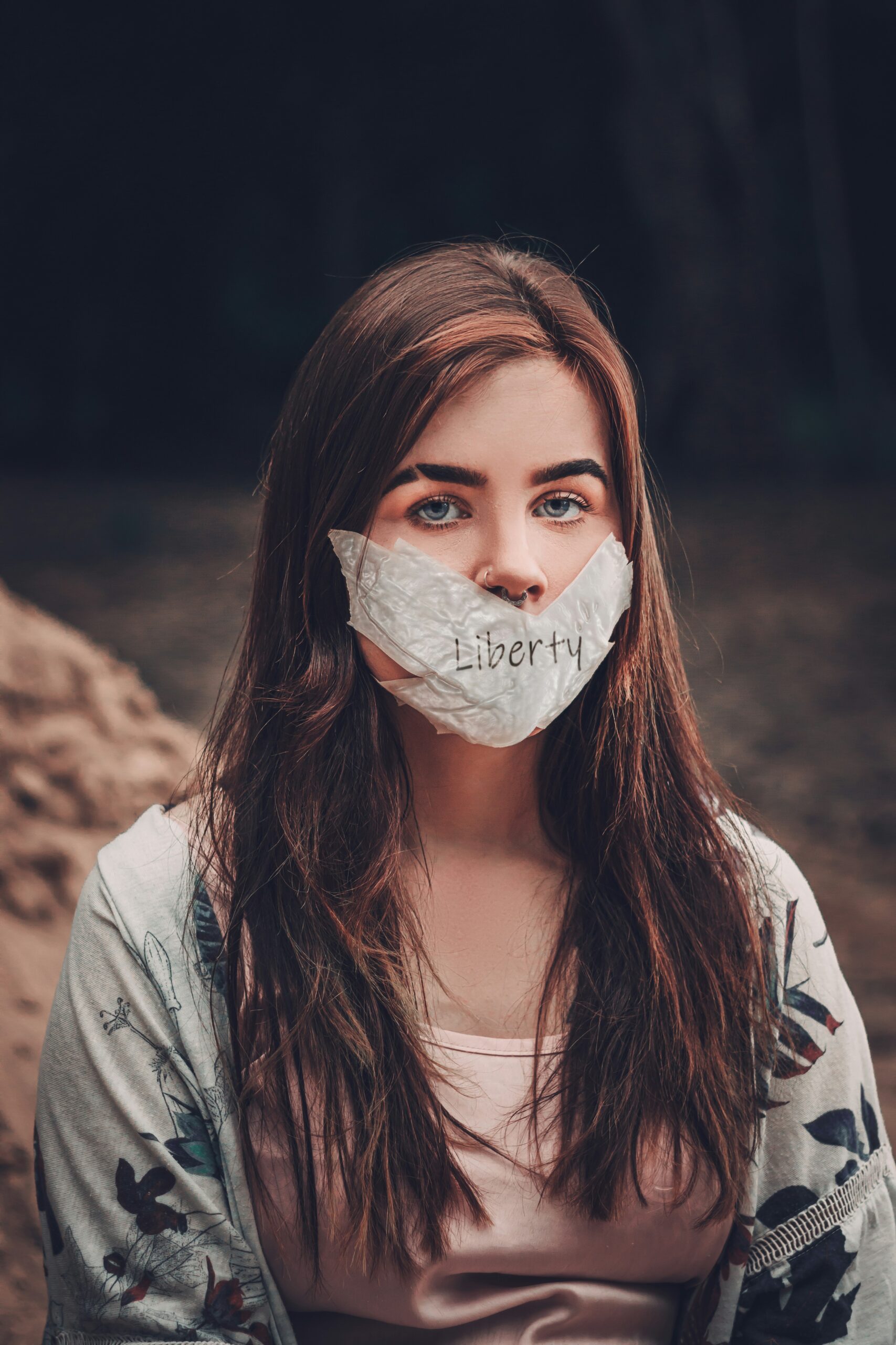 Woman with long brown hair wears a cloth labeled Liberty covering her mouth, sitting outdoors.