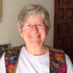 Smiling older woman with short gray hair and glasses, wearing a colorful vest over a white shirt indoors.