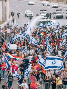 Large crowd marching on a city street, waving Israeli and rainbow flags, with police vehicles nearby.