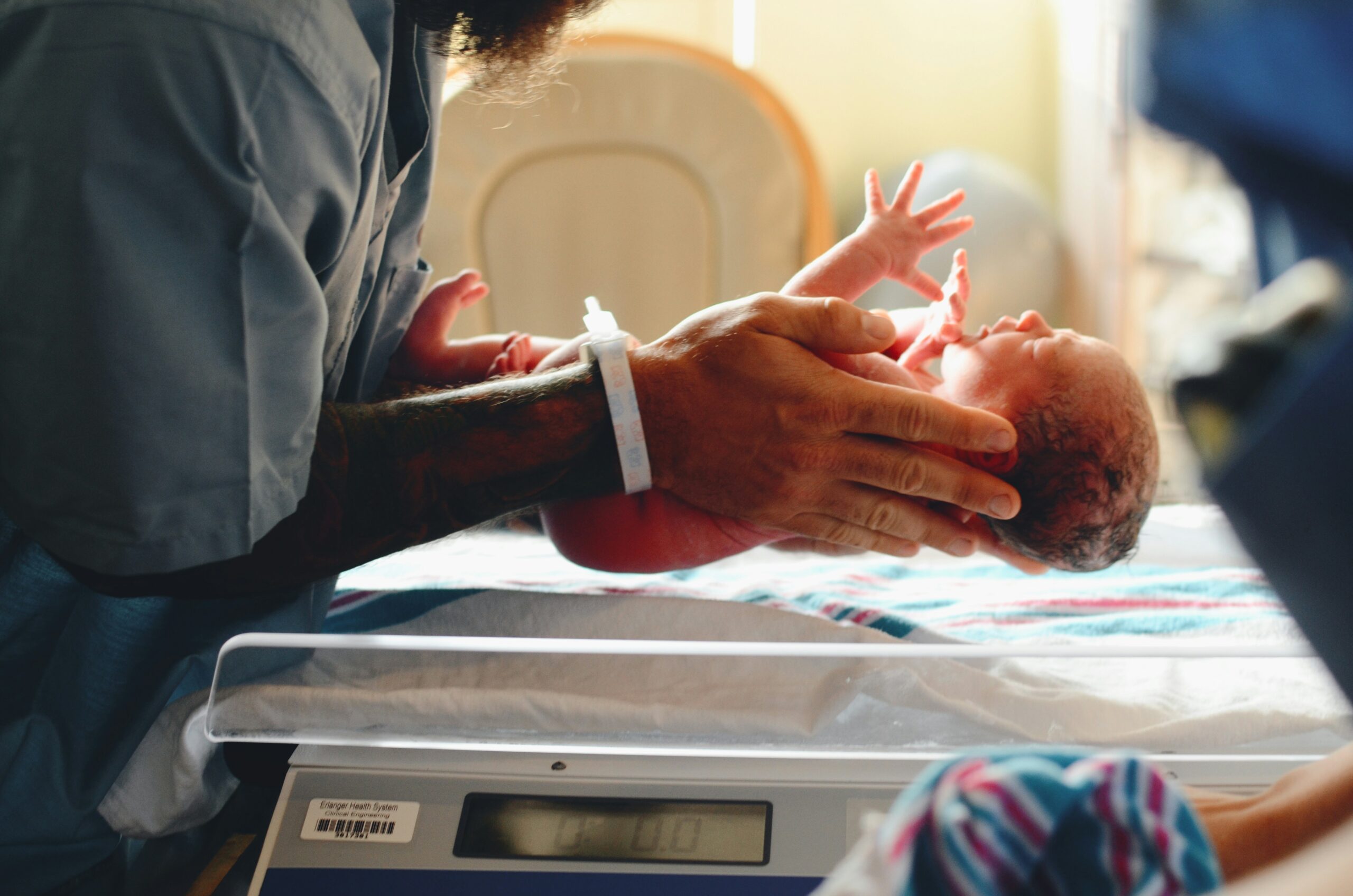 A nurse gently holds a newborn baby above a hospital scale in a brightly lit room.