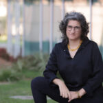 A person with curly gray hair and glasses sits outside near a building, wearing black clothes and a yellow necklace.