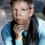 An older woman with short, curly hair and glasses, wearing a blue top and dark scarf, rests her chin on her hand.