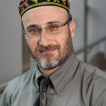 A man with glasses and a patterned cap smiles, wearing a gray shirt and tie against a blurred background.