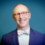 Smiling person in glasses and a bow tie, wearing a blue suit, standing against a teal background.