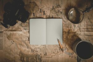 Open notebook, pencil, camera, magnifying glass, and coffee on a vintage map, viewed from above.