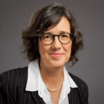 Woman with short brown hair, glasses, and a pinstripe suit smiles against a plain gray background.
