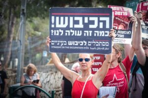 A woman holds a protest sign in Hebrew, English, and Arabic at a demonstration, surrounded by others.