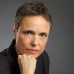 A serious person with short hair in a suit, resting their chin on their hand, looking at the camera.