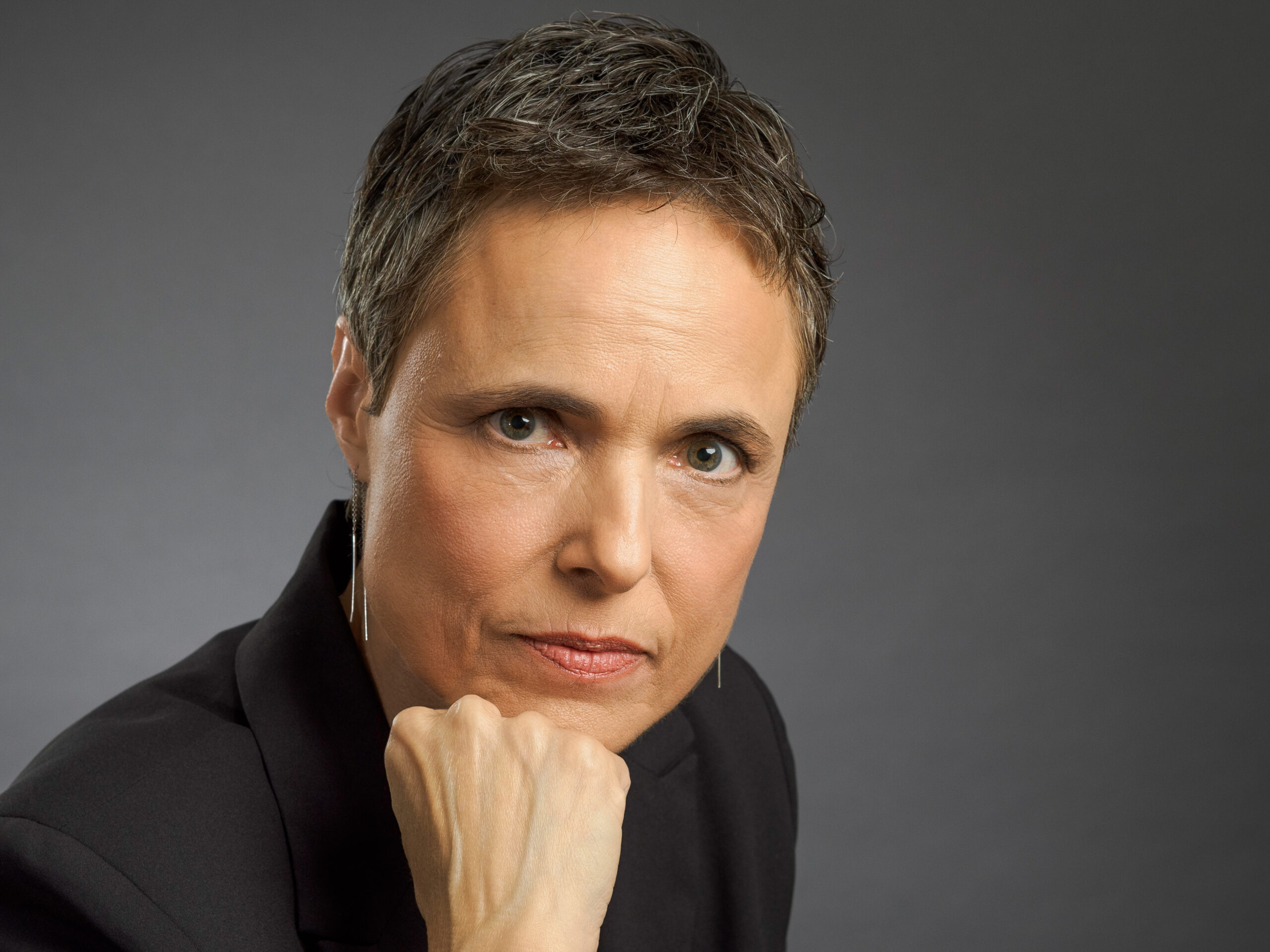 A serious person with short hair in a suit, resting their chin on their hand, looking at the camera.