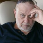 Older man with gray beard and glasses resting on head, sitting and looking intently at the camera.