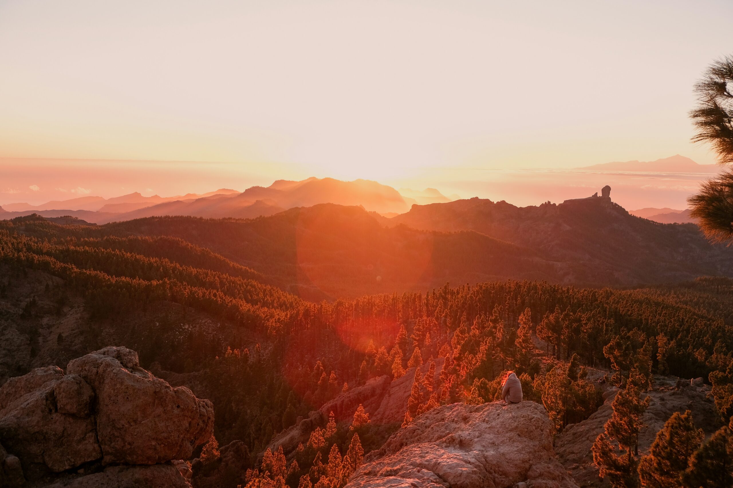 Sunset over a forested mountain landscape with a person sitting on rocks in the foreground.