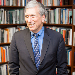 A man in a suit smiles while standing in front of bookshelves filled with books.