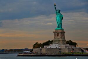 The Statue of Liberty stands on its pedestal with a cloudy sky in the background and water in the foreground.