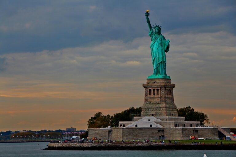 The Statue of Liberty stands on its pedestal with a cloudy sky in the background and water in the foreground.
