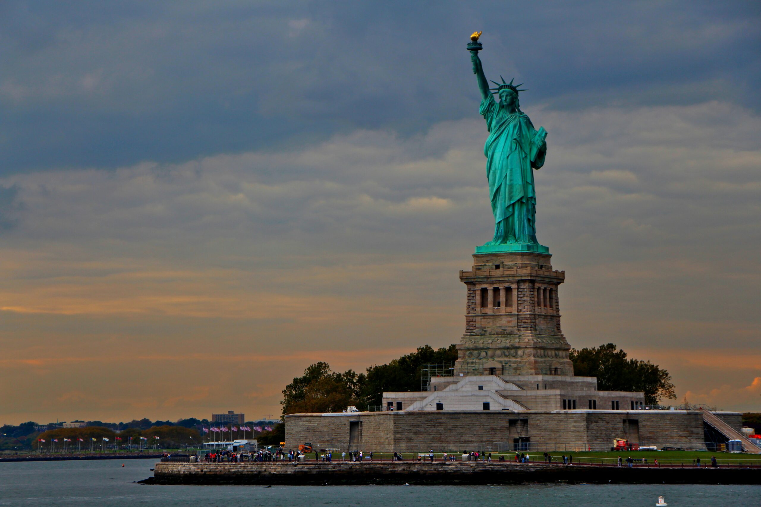 The Statue of Liberty stands on its pedestal with a cloudy sky in the background and water in the foreground.
