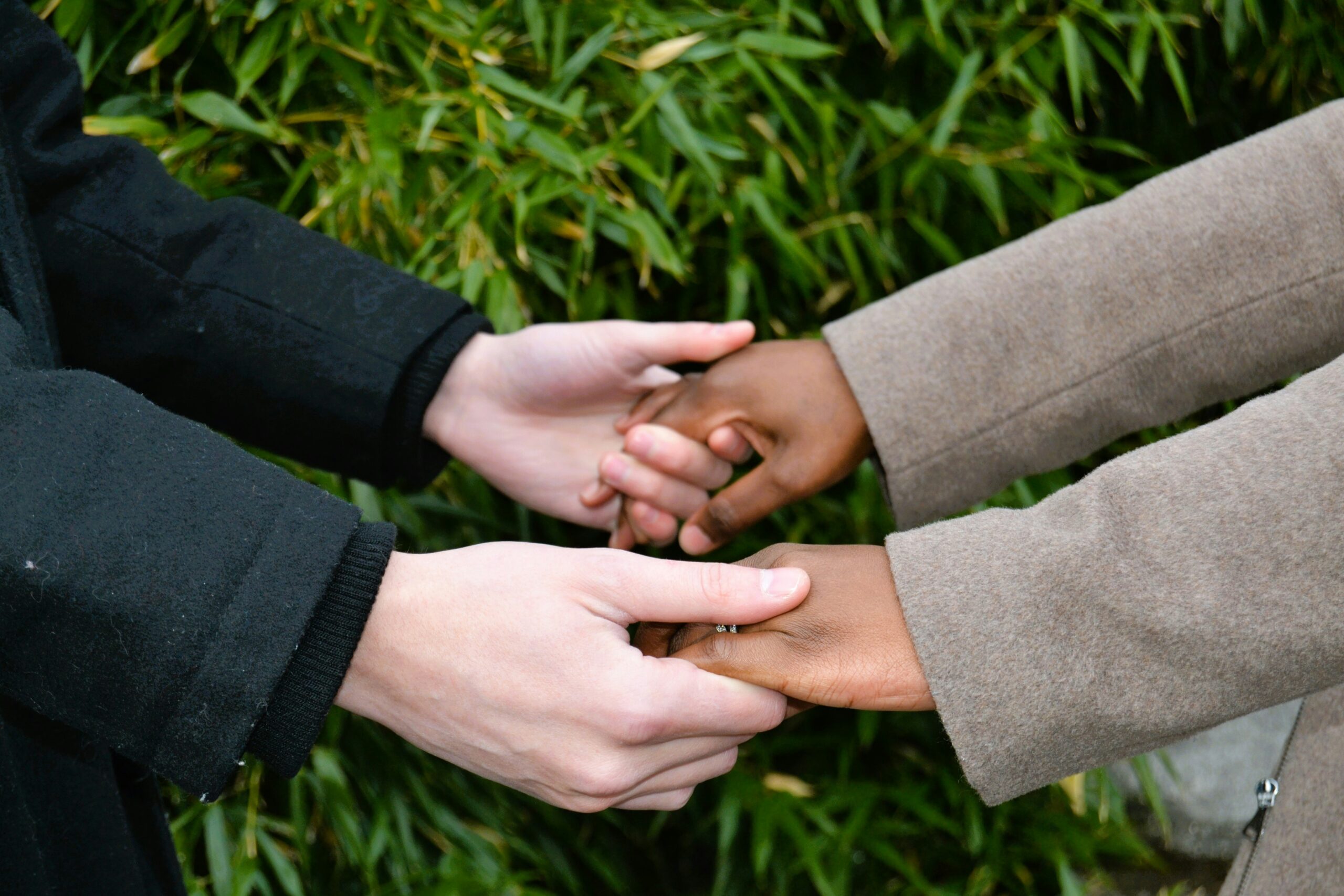 Two people holding hands outdoors, wearing coats, with green leaves in the background.