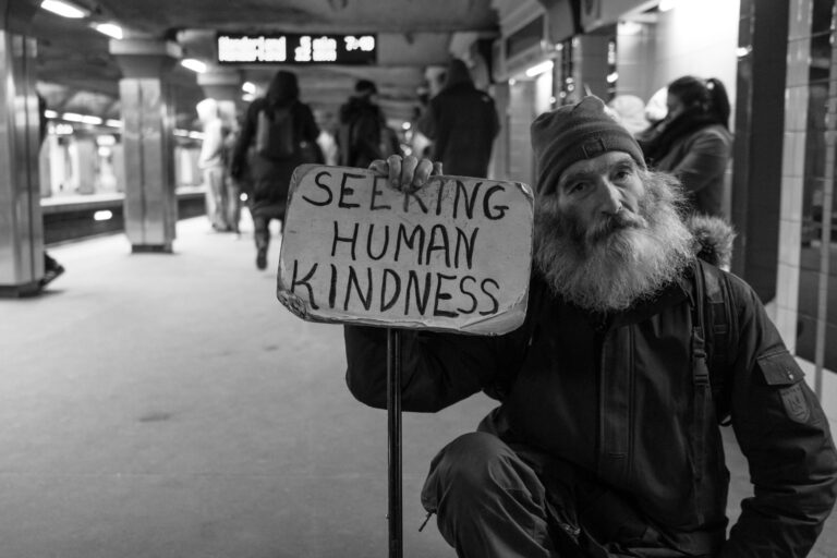 Man in a subway station holds a sign reading Seeking Human Kindness; people walk in the background.