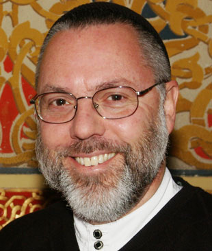 Smiling man with glasses, a gray beard, and a black cap, standing in front of an ornate background.