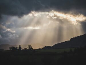 Sunlight rays break through dark clouds over a hilly landscape with silhouetted trees.