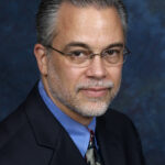 A middle-aged man with glasses, gray hair, and a beard wearing a suit and tie against a blue background.