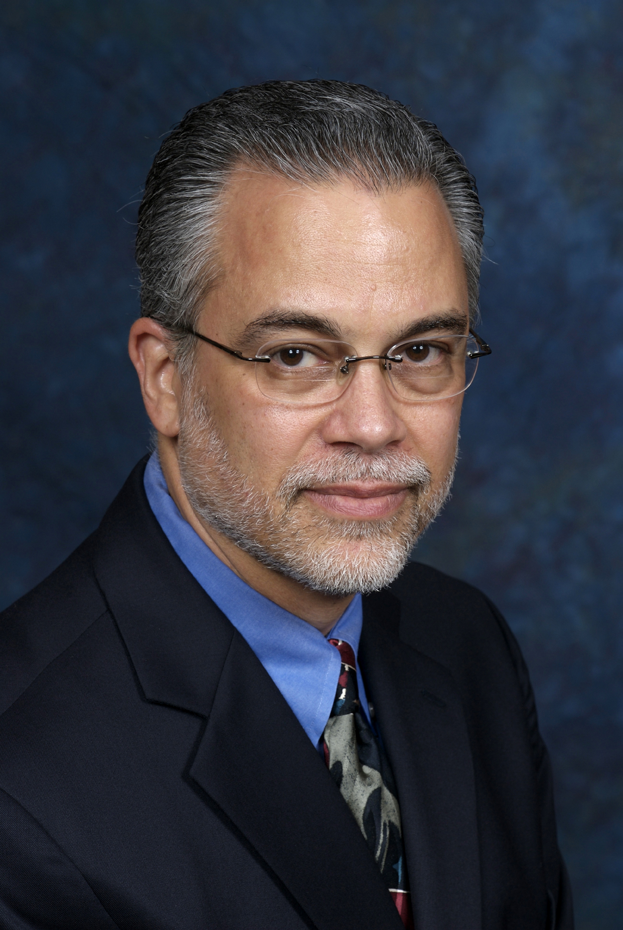 A middle-aged man with glasses, gray hair, and a beard wearing a suit and tie against a blue background.
