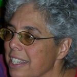 A person with short gray hair, wearing glasses and a red shirt, smiling slightly and looking to the side.