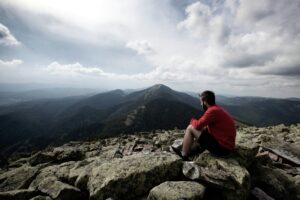 A man in a red jacket sits on rocky ground, gazing at mountain peaks under a cloudy sky.