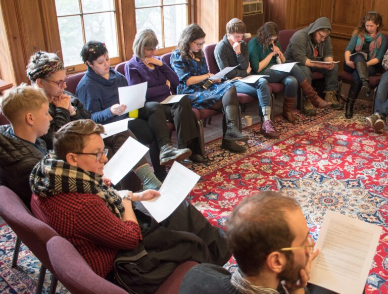 A group of people sit in a circle on chairs, reading papers in a cozy, wood-paneled room with large windows.