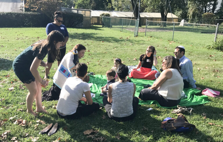 A group of people sit on a blanket outdoors in the sun, listening to a woman holding a colorful chart.