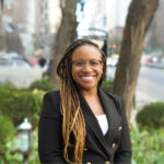 A woman with glasses and long braids smiles outdoors, wearing a black blazer, with greenery and city street behind her.