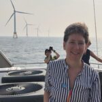 Smiling person on a boat with wind turbines in the ocean and another person taking photos in the background.