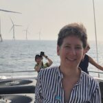 Smiling person on a boat with wind turbines in the ocean and another person taking photos in the background.