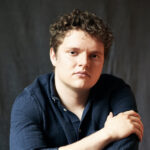 Person with short curly hair sits at a table, arms crossed, wearing a dark blue shirt, gazing at the camera.