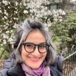 Woman with glasses smiles in front of blooming white flowers and greenery, wearing a scarf and jacket outdoors.