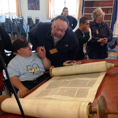 A man helps a boy read from an open Torah scroll in a room with other people and stacked chairs.