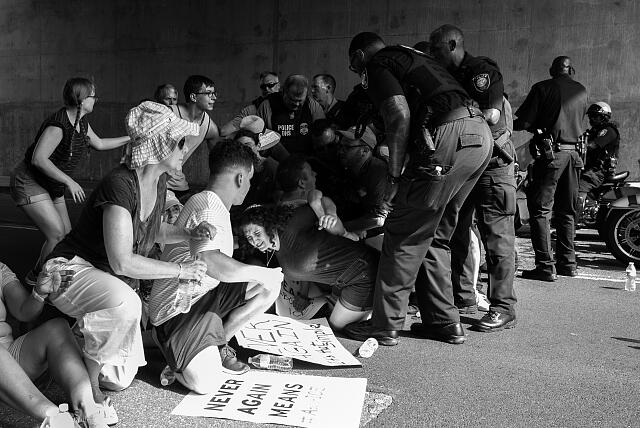 Protesters sitting on the ground as police officers detain someone; protest signs are visible.