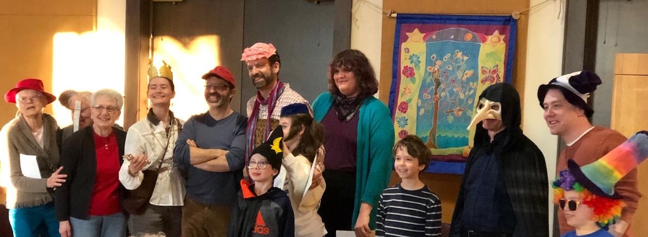 A group of people, adults and children, dressed in colorful costumes and hats, posing indoors and smiling.