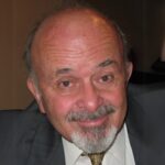 Older man with a bald head, gray beard, and mustache, wearing a suit and tie, smiling at the camera.