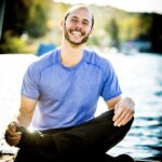 Smiling man in a blue shirt sits cross-legged on a dock by the water on a sunny day.
