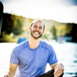 Smiling man in a blue shirt sits cross-legged on a dock by the water on a sunny day.