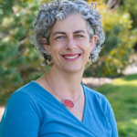 A woman with short curly gray hair and a blue top smiles outdoors with greenery in the background.