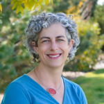 A woman with short curly gray hair and a blue top smiles outdoors with greenery in the background.