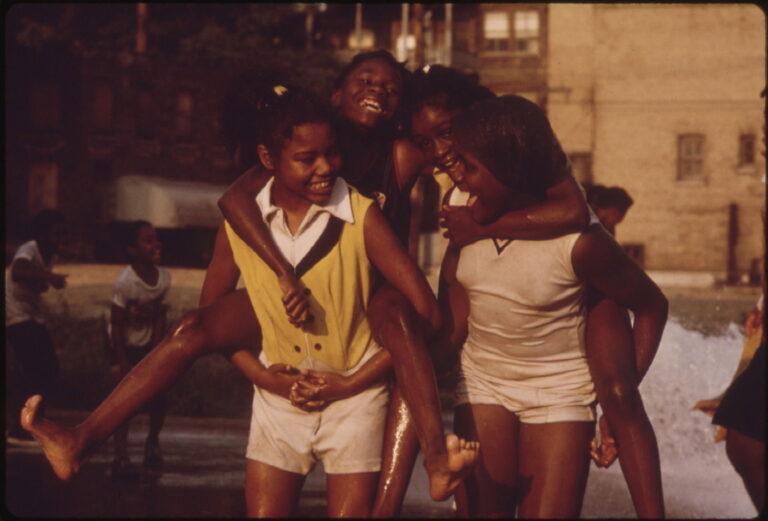 Four girls play and give piggyback rides at a park on a sunny day, smiling and laughing together.