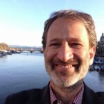 A man with a beard smiling by a river with boats and city buildings in the background on a sunny day.