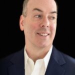A man in a navy suit and white shirt looks to the side against a black background.