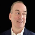 A man in a navy suit and white shirt looks to the side against a black background.