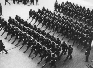 A group of uniformed soldiers march in formation on a paved street, viewed from above.
