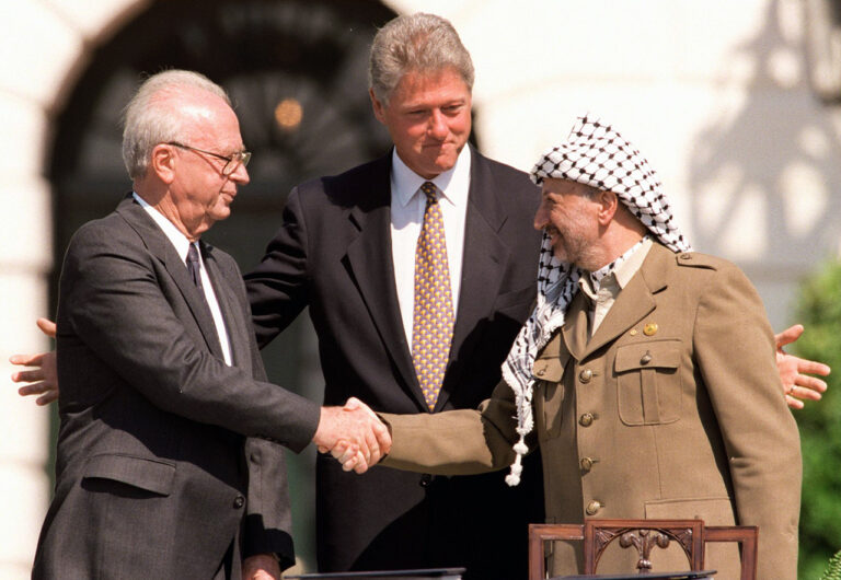 Three men in suits and uniform shake hands outdoors, with one man standing between them, smiling and guiding the handshake.